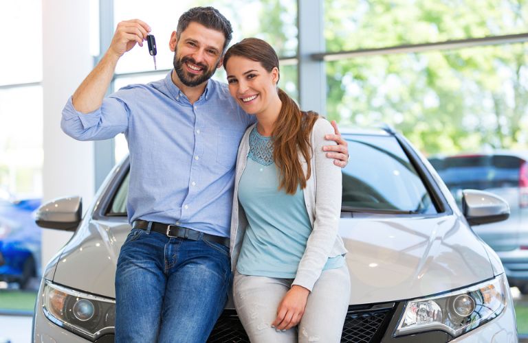 a man and a woman holding a black car key posing in front of a car